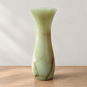 May include: A tall, slender vase crafted from smooth, pale green stone with a flared rim. The surface features natural, irregular brown veining that runs vertically along the body, creating a unique pattern against the translucent light green base.