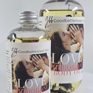 Könnte beinhalten: Zwei klare Glasflaschen mit Körperöl und silbernen Verschlüssen. Die Etiketten zeigen ein Foto eines sich küssenden Paares und die Worte "LOVE PHEROMONE BODY OIL". Das Öl hat eine hellgelbe Farbe.