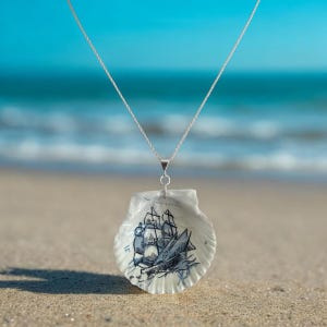 May include: A silver-toned chain necklace featuring a white scallop shell pendant. The interior of the shell is decorated with a detailed black ink illustration of a multi-masted sailing ship with visible sails and rigging.