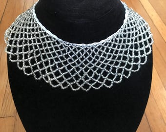 May include: A silver beaded necklace with a net-like design. The necklace is displayed on a black mannequin head against a wooden background. The beads are small and round, creating a delicate, openwork pattern.