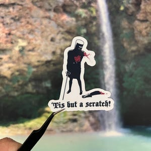 May include: A sticker featuring a black knight with a red heart and blood splatter, holding a cane, with a severed arm on the ground. The text reads, "'Tis but a scratch!"