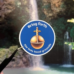 May include: A round sticker with a blue border and the text "Bring forth" at the top. The center features a gold-colored orb with a cross on top, and the text "The holy hand grenade" at the bottom. The sticker is held by tweezers.