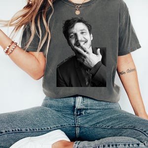 Op de afbeelding: Donkergrijs T-shirt met een zwart-wit afbeelding van een lachende persoon. De afbeelding heeft de tekst "PEDRO PASCAL" erop gedrukt. Het woord "PEPPER" staat rechtsboven op het shirt.