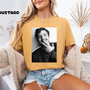 Op de afbeelding: Mosterdkleurig T-shirt met een zwart-wit afbeelding van een lachende persoon. De afbeelding heeft de tekst "PEDRO PASCAL" verticaal afgedrukt. Het woord "MUSTARD" is in zwarte blokletters boven de afbeelding gedrukt.