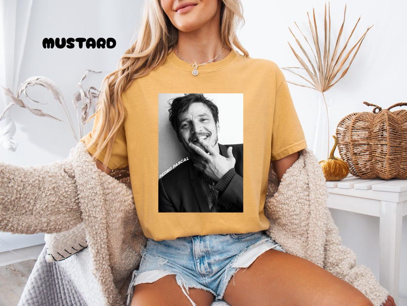 Op de afbeelding: Mosterdkleurig T-shirt met een zwart-wit afbeelding van een lachende persoon. De afbeelding heeft de tekst "PEDRO PASCAL" verticaal afgedrukt. Het woord "MUSTARD" is in zwarte blokletters boven de afbeelding gedrukt.