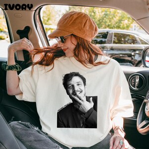 Op de afbeelding: Ivoorkleurige sweatshirt met een zwart-wit afbeelding van Pedro Pascal. De afbeelding is gecentreerd op de voorkant van de trui. De tekst "PEDRO PASCAL" is onder de afbeelding gedrukt. De persoon op de foto draagt een bruine corduroy hoed en een zonnebril.