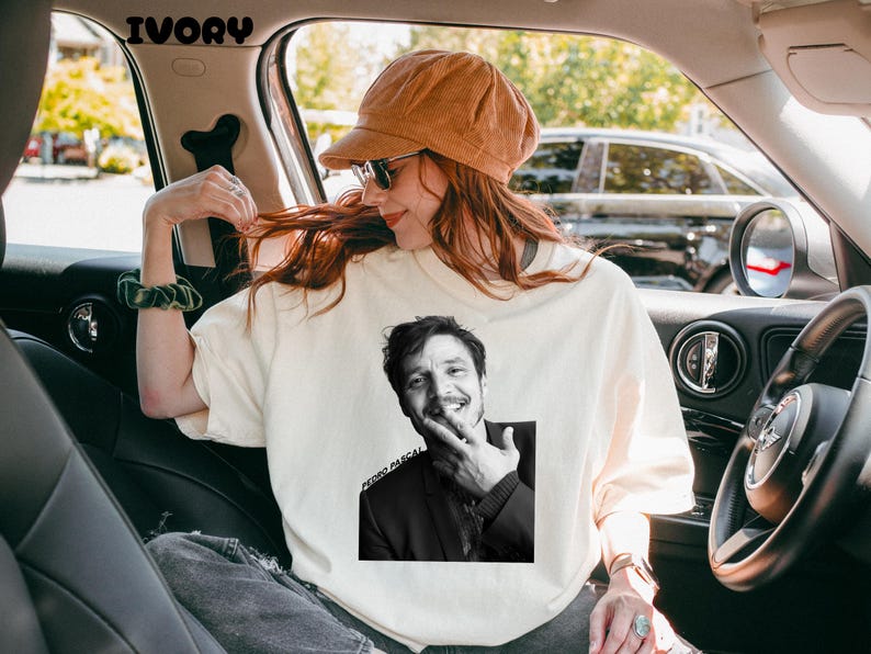 Op de afbeelding: Ivoorkleurige sweatshirt met een zwart-wit afbeelding van Pedro Pascal. De afbeelding is gecentreerd op de voorkant van de trui. De tekst "PEDRO PASCAL" is onder de afbeelding gedrukt. De persoon op de foto draagt een bruine corduroy hoed en een zonnebril.