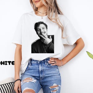 Op de afbeelding: Wit T-shirt met een zwart-wit afbeelding van Pedro Pascal. De afbeelding toont Pascal die lacht en zijn gezicht aanraakt. De tekst "PEDRO PASCAL" is eronder gedrukt. Het woord "WHITE" staat links onderin in het zwart.