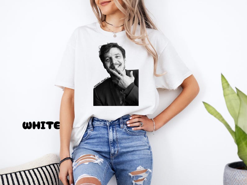 Op de afbeelding: Wit T-shirt met een zwart-wit afbeelding van Pedro Pascal. De afbeelding toont Pascal die lacht en zijn gezicht aanraakt. De tekst "PEDRO PASCAL" is eronder gedrukt. Het woord "WHITE" staat links onderin in het zwart.