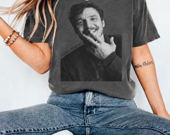 Könnte beinhalten: Ein anthrazitfarbenes Kurzarm-T-Shirt mit Rundhalsausschnitt und einem schwarz-weißen Fotodruck von Pedro Pascal. Die Grafik zeigt ein Porträt des Schauspielers, wobei der Text "PEDRO PASCAL" diagonal in der unteren linken Ecke gedruckt ist.