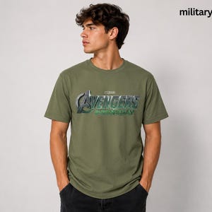 K&ouml;nnte beinhalten: Ein milit&auml;rgr&uuml;nes Kurzarm-T-Shirt mit dem metallisch silbernen und gr&uuml;nen Avengers Endgame-Logo auf der Brust. Das Shirt verf&uuml;gt &uuml;ber einen Rundhalsausschnitt und eine Standardpassform, die das ikonische Film-Branding als Grafikdesign zeigt.