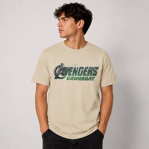 K&ouml;nnte beinhalten: Ein sandfarbenes Kurzarm-T-Shirt mit dem auf die Brust gedruckten Text Avengers Doomsday. Die Grafik enth&auml;lt ein gro&szlig;es stilisiertes A und das Wort Avengers in einem metallischen Gr&uuml;n- und Silberverlauf, mit Doomsday darunter.