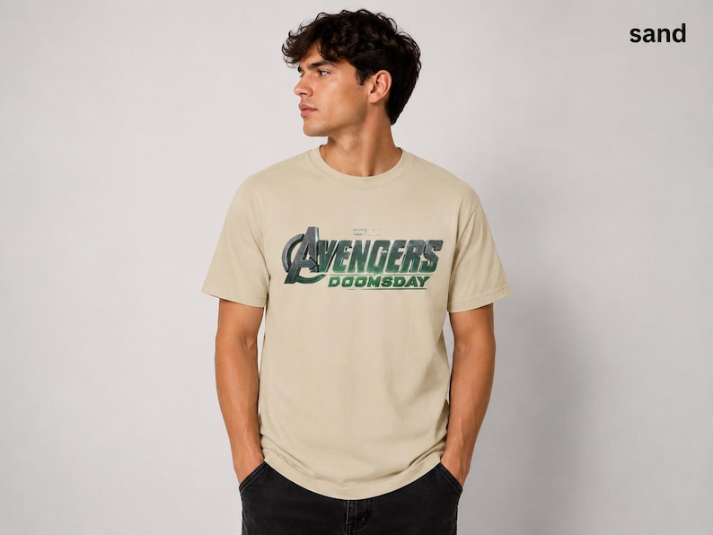 K&ouml;nnte beinhalten: Ein sandfarbenes Kurzarm-T-Shirt mit dem auf die Brust gedruckten Text Avengers Doomsday. Die Grafik enth&auml;lt ein gro&szlig;es stilisiertes A und das Wort Avengers in einem metallischen Gr&uuml;n- und Silberverlauf, mit Doomsday darunter.