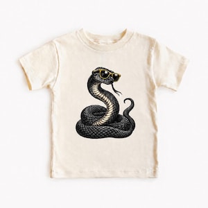 Peut inclure: Un t-shirt à manches courtes de couleur crème arborant une illustration graphique d'un serpent noir enroulé portant des lunettes de soleil à monture dorée. Le serpent présente un motif d'écailles détaillé et est centré sur le t-shirt.
