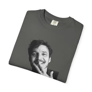 Op de afbeelding: Donkergrijs t-shirt met een zwart-wit afbeelding van een lachende persoon. De persoon raakt zijn gezicht aan met zijn vingers. Het t-shirt heeft een ronde hals en een label met de tekst "Comfort Colors".