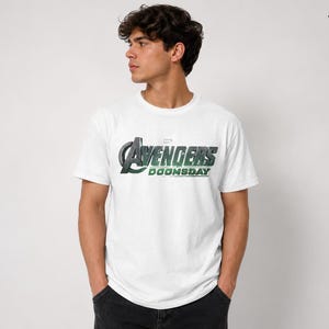 K&ouml;nnte beinhalten: Ein wei&szlig;es T-Shirt mit kurzen &Auml;rmeln und dem auf die Brust gedruckten Avengers Doomsday-Logo. Die Grafik zeigt das ikonische Avengers A-Symbol in metallischer Optik mit gr&uuml;nen Akzenten sowie den Schriftzug Doomsday in Fettdruck.