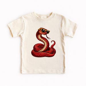 Op de afbeelding: Een crèmekleurig T-shirt met korte mouwen en een grafische print van een opgerolde rode slang met een zonnebril met geel montuur. De slang is afgebeeld met een textuur van schubben, een zichtbare tong en een gekrulde staart op de borst.