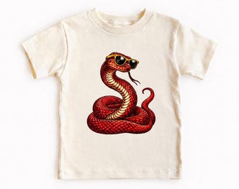 Könnte beinhalten: Ein cremefarbenes Kurzarm-T-Shirt mit einem Grafikdruck einer geringelten roten Schlange, die eine Sonnenbrille mit gelbem Rahmen trägt. Die Schlange ist mit einem strukturierten Schuppenmuster, einer sichtbaren Zunge und einem geschwungenen Schwanz dargestellt.