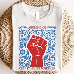 May include: White t-shirt featuring a red fist graphic within a blue floral design. The text "ABOLISH ICE" is above the fist, and "KEEP FAMILIES TOGETHER" is below. The shirt is displayed on a woven, circular surface.