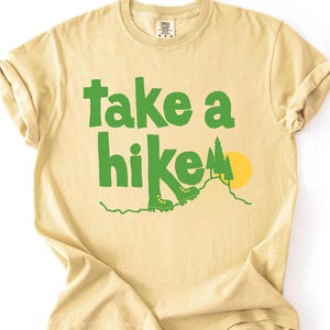 May include: A light yellow t-shirt with the words "take a hike" in green block letters. The design includes green hiking boots, a mountain, and a yellow sun. The shirt is made of a soft material and has rolled-up sleeves.