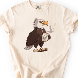 Puede incluir: Camiseta color crema con un &aacute;guila de dibujos animados sosteniendo una taza con la frase "IS HE DEAD YET?". El &aacute;guila es marr&oacute;n, blanca y gris, con pico y garras amarillas. La camiseta tiene las mangas enrolladas. El dise&ntilde;o est&aacute; impreso.
