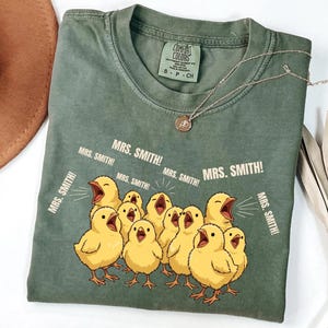 May include: A sage green t-shirt with a graphic of yellow chicks yelling "MRS. SMITH!". The shirt has a crew neck and short sleeves. A silver necklace with a pendant is draped on the shirt.