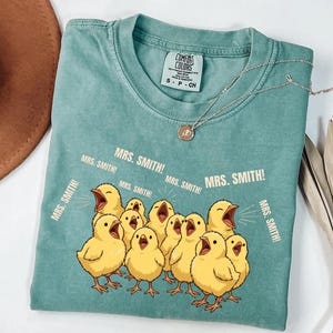 May include: A teal Comfort Colors t-shirt featuring a graphic of yellow chicks with open beaks, and the text "MRS. SMITH!". A silver necklace with a pendant is draped across the shirt. The shirt is folded on a white surface.