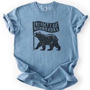 May include: A blue t-shirt with a black bear holding a flag that reads 'Protect Our National Parks'.