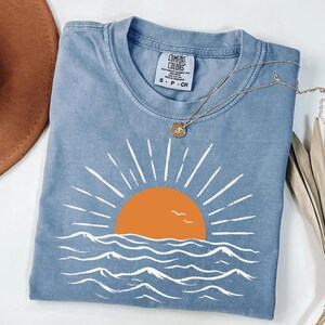 May include: A blue short-sleeve t-shirt featuring a graphic of an orange sun rising over white stylized ocean waves. The shirt has a Comfort Colors label at the neck, which reads Comfort Colors, Heavyweight Garment Dye, 100% Cotton, Made in Honduras, S-P-CH.