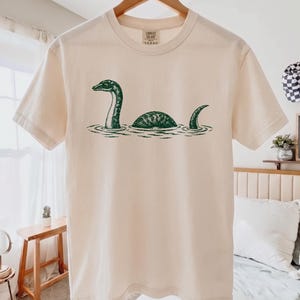 May include: A cream-colored short-sleeve t-shirt featuring a dark green graphic print of the Loch Ness monster swimming in water. The design shows the creature's head, back, and tail emerging from the surface, centered on the chest of the shirt.