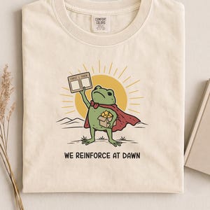 May include: A cream-colored t-shirt featuring a graphic of a green frog wearing a red cape. The frog holds a sign labeled FIRST THEN and a box of yellow stars. Behind the frog is a yellow sun. The text WE REINFORCE AT DAWN is printed below.