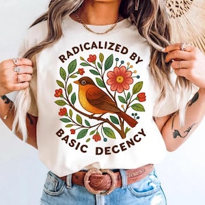 May include: Cream-colored t-shirt with the text "RADICALIZED BY BASIC DECENCY" in a circular design. The design features a brown bird perched on a branch surrounded by red flowers and green leaves.