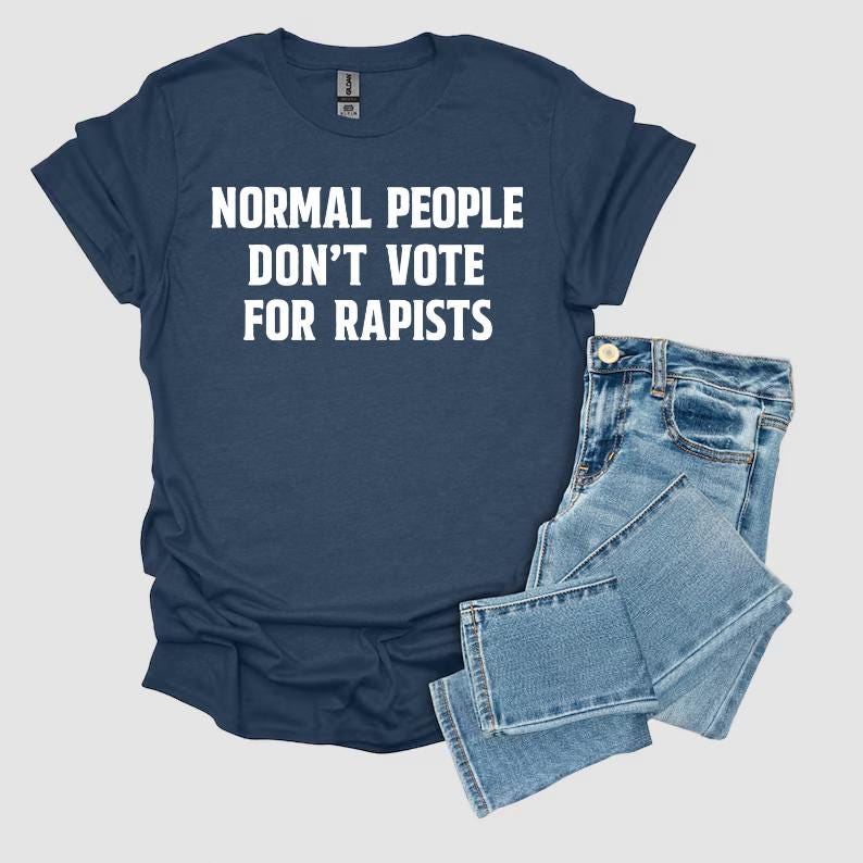 May include: A navy blue t-shirt with the text "NORMAL PEOPLE DON'T VOTE FOR RAPISTS" in white. The shirt is paired with light blue denim jeans.