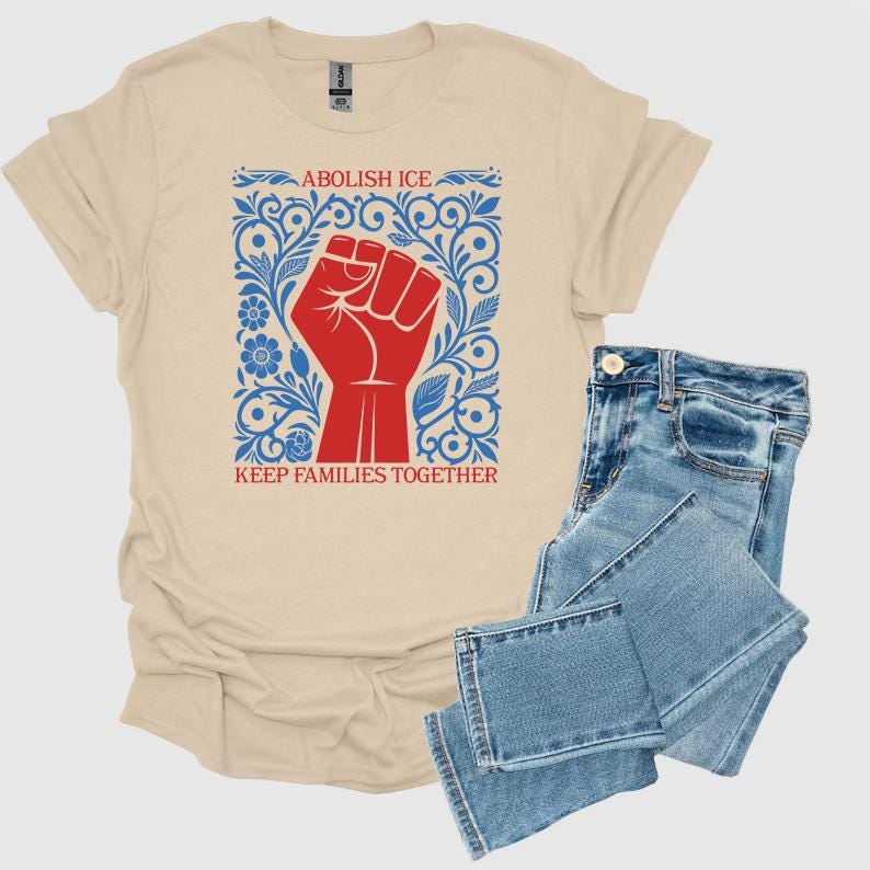 May include: A beige t-shirt with a red fist graphic, blue floral accents, and the text "ABOLISH ICE" and "KEEP FAMILIES TOGETHER." The shirt is paired with blue denim jeans.