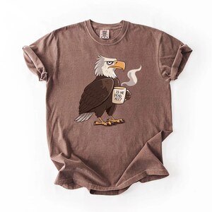 Puede incluir: Camiseta marr&oacute;n con un dise&ntilde;o de &aacute;guila calva de dibujos animados sosteniendo una taza de caf&eacute;. El &aacute;guila tiene plumas blancas en la cabeza y un pico y patas amarillas. La taza dice "IS HE DEAD YET?" en texto negro.