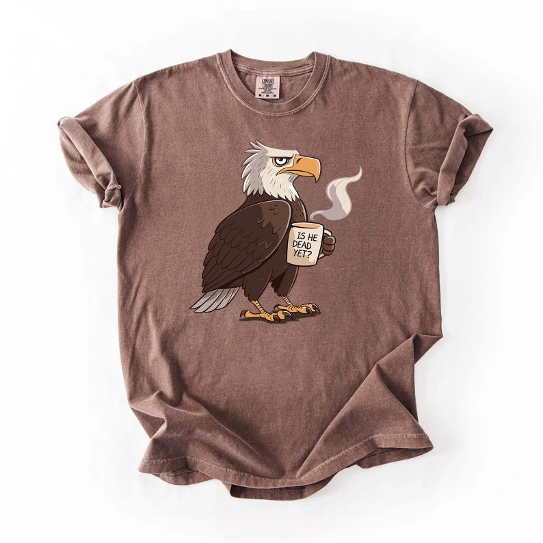 Puede incluir: Camiseta marr&oacute;n con un dise&ntilde;o de &aacute;guila calva de dibujos animados sosteniendo una taza de caf&eacute;. El &aacute;guila tiene plumas blancas en la cabeza y un pico y patas amarillas. La taza dice "IS HE DEAD YET?" en texto negro.