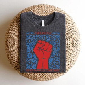 May include: Dark gray t-shirt with a red fist graphic inside a blue floral frame. The text "ABOLISH ICE" and "KEEP FAMILIES TOGETHER" are in red. The shirt is folded on a woven, circular surface.