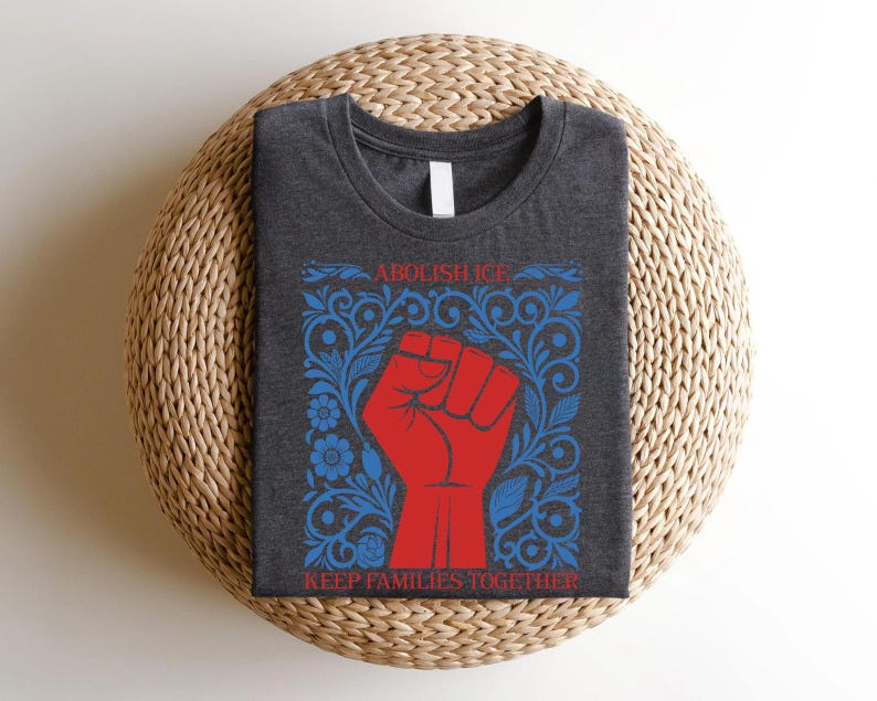 May include: Dark gray t-shirt with a red fist graphic inside a blue floral frame. The text "ABOLISH ICE" and "KEEP FAMILIES TOGETHER" are in red. The shirt is folded on a woven, circular surface.
