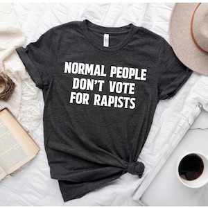 May include: Dark gray t-shirt with the white text "NORMAL PEOPLE DON'T VOTE FOR RAPISTS." The shirt is knotted at the bottom. A beige hat and a cup of coffee are in the background.