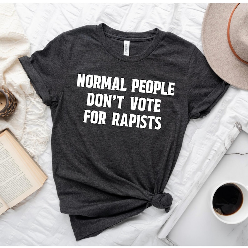 May include: Dark gray t-shirt with the white text "NORMAL PEOPLE DON'T VOTE FOR RAPISTS." The shirt is knotted at the bottom. A beige hat and a cup of coffee are in the background.