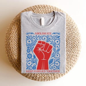 May include: A heather gray t-shirt featuring a red fist graphic within a blue floral design. The text "ABOLISH ICE" and "KEEP FAMILIES TOGETHER" are printed above and below the fist. The shirt is displayed on a woven, round surface.