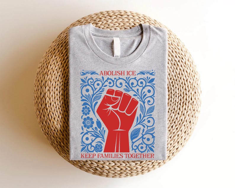 May include: A heather gray t-shirt featuring a red fist graphic within a blue floral design. The text "ABOLISH ICE" and "KEEP FAMILIES TOGETHER" are printed above and below the fist. The shirt is displayed on a woven, round surface.
