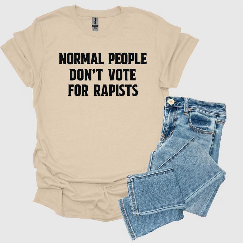 May include: A beige t-shirt with the text "NORMAL PEOPLE DON'T VOTE FOR RAPISTS" in black, paired with blue denim jeans. The t-shirt has short sleeves and a crew neck. The jeans are folded.
