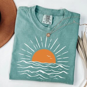 May include: A sage green short-sleeve t-shirt featuring a graphic of an orange sun setting over white stylized ocean waves. The shirt has a crew neckline and a Comfort Colors brand label on the inside collar. A thin gold chain necklace rests on top.