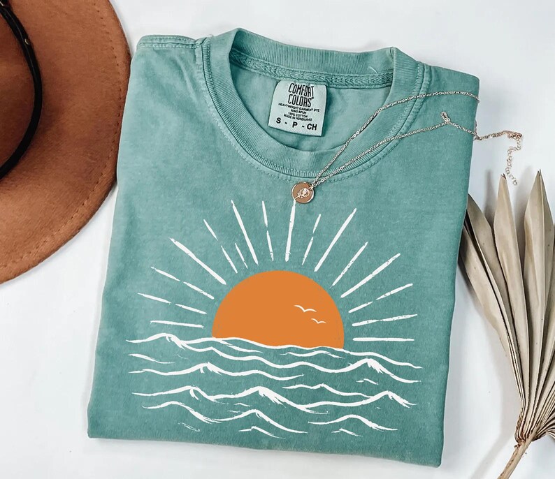 May include: A sage green short-sleeve t-shirt featuring a graphic of an orange sun setting over white stylized ocean waves. The shirt has a crew neckline and a Comfort Colors brand label on the inside collar. A thin gold chain necklace rests on top.
