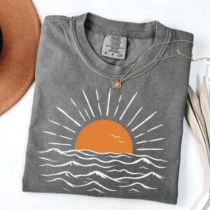 May include: A charcoal gray short-sleeve t-shirt featuring a graphic of an orange sun rising over white stylized ocean waves. The shirt has a crew neckline and a Comfort Colors brand label printed on the inside collar, indicating a size small.