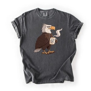 Puede incluir: Camiseta gris oscuro con un &aacute;guila de dibujos animados sosteniendo una taza humeante. El &aacute;guila tiene plumas blancas, alas marrones y un pico y patas naranjas. La taza dice "IS HE DEAD YET?" en texto negro. La camiseta tiene las mangas enrolladas.
