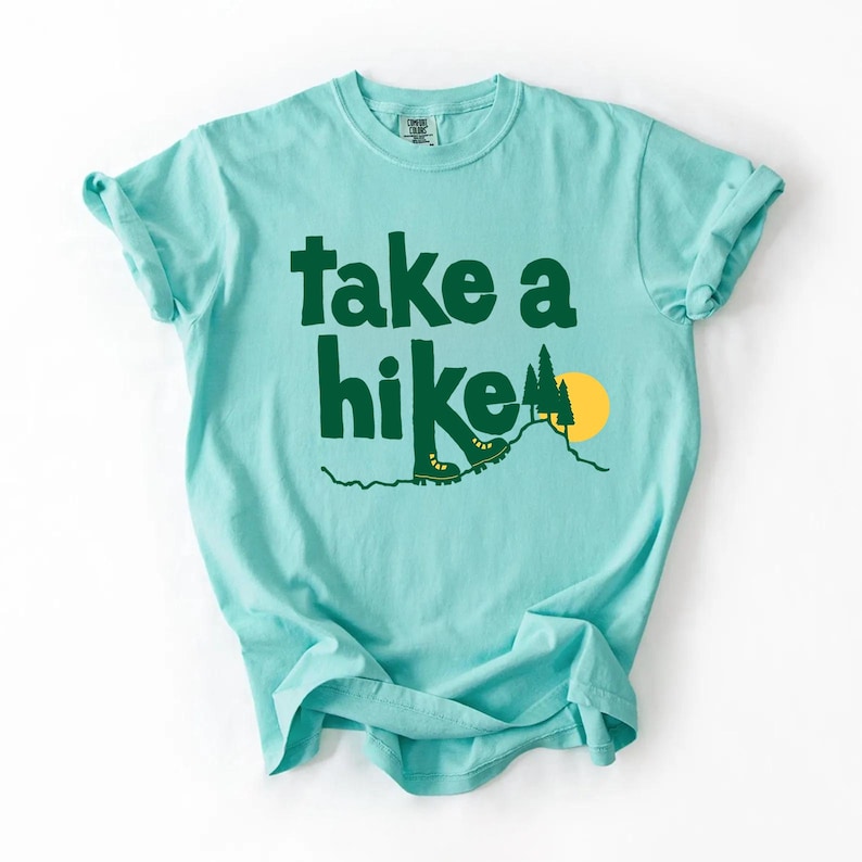 May include: A light blue t-shirt with the words "take a hike" in dark green block letters. The design includes a mountain scene with hiking boots, a tree, and a yellow sun. The shirt is made of a soft material.