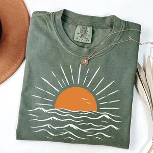 May include: An olive green t-shirt featuring a graphic of an orange sun rising over white stylized ocean waves with two small birds. The shirt has a crew neckline and a Comfort Colors brand label on the inside collar indicating size small.