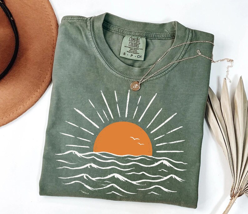 May include: An olive green t-shirt featuring a graphic of an orange sun rising over white stylized ocean waves with two small birds. The shirt has a crew neckline and a Comfort Colors brand label on the inside collar indicating size small.